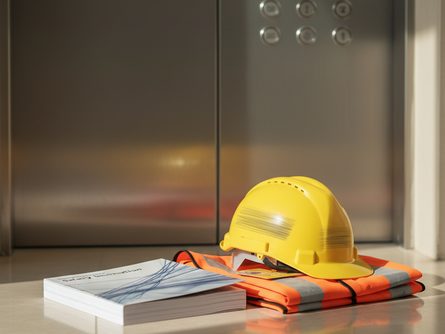 엘리베이터 버튼 패널 앞에 안전 안내 책자와 헬멧, 안전 조끼가 놓인 모습